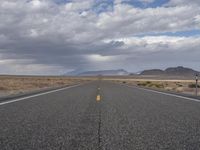 an empty highway in the middle of nowhere with mountains in the background and a cloudy sky overhead