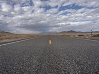 an empty highway in the middle of nowhere with mountains in the background and a cloudy sky overhead