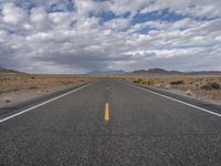 an empty highway in the middle of nowhere with mountains in the background and a cloudy sky overhead