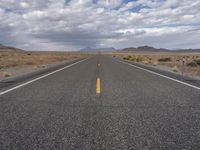 an empty highway in the middle of nowhere with mountains in the background and a cloudy sky overhead