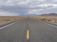 an empty highway in the middle of nowhere with mountains in the background and a cloudy sky overhead