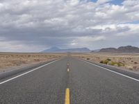 an empty highway in the middle of nowhere with mountains in the background and a cloudy sky overhead