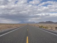 an empty highway in the middle of nowhere with mountains in the background and a cloudy sky overhead