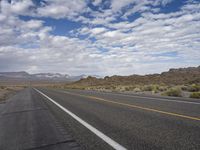 an empty highway in the desert with mountains and sky above it and clouds overhead on a bright, cloudy day