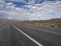 an empty highway in the desert with mountains and sky above it and clouds overhead on a bright, cloudy day