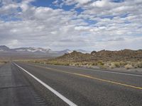 an empty highway in the desert with mountains and sky above it and clouds overhead on a bright, cloudy day