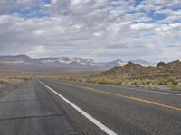 an empty highway in the desert with mountains and sky above it and clouds overhead on a bright, cloudy day