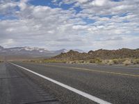 an empty highway in the desert with mountains and sky above it and clouds overhead on a bright, cloudy day