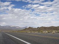 an empty highway in the desert with mountains and sky above it and clouds overhead on a bright, cloudy day