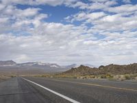 an empty highway in the desert with mountains and sky above it and clouds overhead on a bright, cloudy day