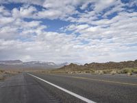 an empty highway in the desert with mountains and sky above it and clouds overhead on a bright, cloudy day