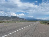 an asphalt road with mountains in the distance, with white clouds in the sky and light yellow lines on the ground