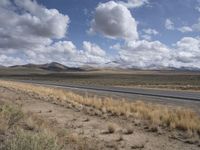 a lone, empty highway surrounded by barren land and mountains at the horizon with puffy clouds above