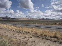 a lone, empty highway surrounded by barren land and mountains at the horizon with puffy clouds above