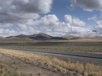 a lone, empty highway surrounded by barren land and mountains at the horizon with puffy clouds above