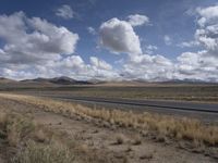 a lone, empty highway surrounded by barren land and mountains at the horizon with puffy clouds above