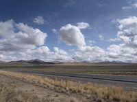 a lone, empty highway surrounded by barren land and mountains at the horizon with puffy clouds above