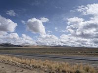 a lone, empty highway surrounded by barren land and mountains at the horizon with puffy clouds above