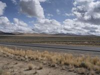 a lone, empty highway surrounded by barren land and mountains at the horizon with puffy clouds above