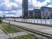 the train tracks are next to a bridge and grass near by water and buildings with a clock tower