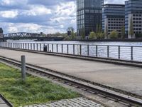 the train tracks are next to a bridge and grass near by water and buildings with a clock tower