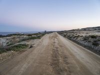 a dirt road with some hills behind it and the ocean in the distance and the sky