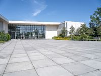 a photo of the exterior of a large white building with many windows, trees, bushes and stone flooring