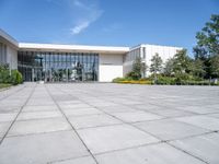 a photo of the exterior of a large white building with many windows, trees, bushes and stone flooring