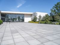 a photo of the exterior of a large white building with many windows, trees, bushes and stone flooring