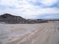 a dirt road next to a bunch of rocks and gravel with a train bridge in the background