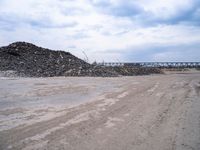 a dirt road next to a bunch of rocks and gravel with a train bridge in the background