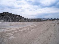 a dirt road next to a bunch of rocks and gravel with a train bridge in the background