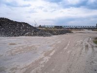 a dirt road next to a bunch of rocks and gravel with a train bridge in the background