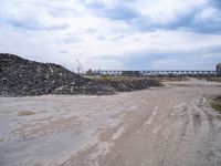 a dirt road next to a bunch of rocks and gravel with a train bridge in the background