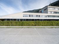 a paved parking lot outside a building and a large hedge lined hillside behind it to the right