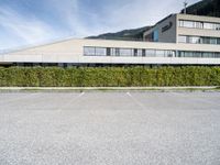 a paved parking lot outside a building and a large hedge lined hillside behind it to the right