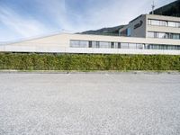 a paved parking lot outside a building and a large hedge lined hillside behind it to the right