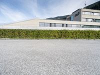 a paved parking lot outside a building and a large hedge lined hillside behind it to the right