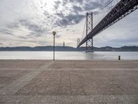 the big bridge is seen across the water from the sea shore by the road and a bench under it