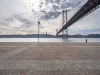 the big bridge is seen across the water from the sea shore by the road and a bench under it