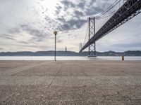 the big bridge is seen across the water from the sea shore by the road and a bench under it