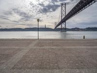 the big bridge is seen across the water from the sea shore by the road and a bench under it