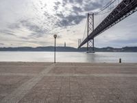 the big bridge is seen across the water from the sea shore by the road and a bench under it