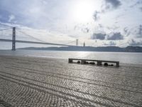 some benches are sitting on the beach near the water, and there is the bridge in the distance