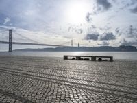 some benches are sitting on the beach near the water, and there is the bridge in the distance