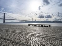 some benches are sitting on the beach near the water, and there is the bridge in the distance