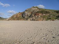 an empty beach with footprints and rock formations in the background, and in the foreground is a steep, grassy hillside