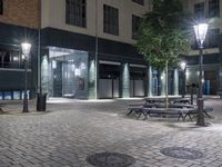 several benches on a brick floor in front of an apartment building at night with some street lights