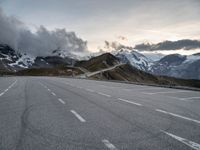 empty road surrounded by large mountains under cloudy skies over them and the snow covered peaks above