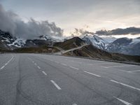 empty road surrounded by large mountains under cloudy skies over them and the snow covered peaks above